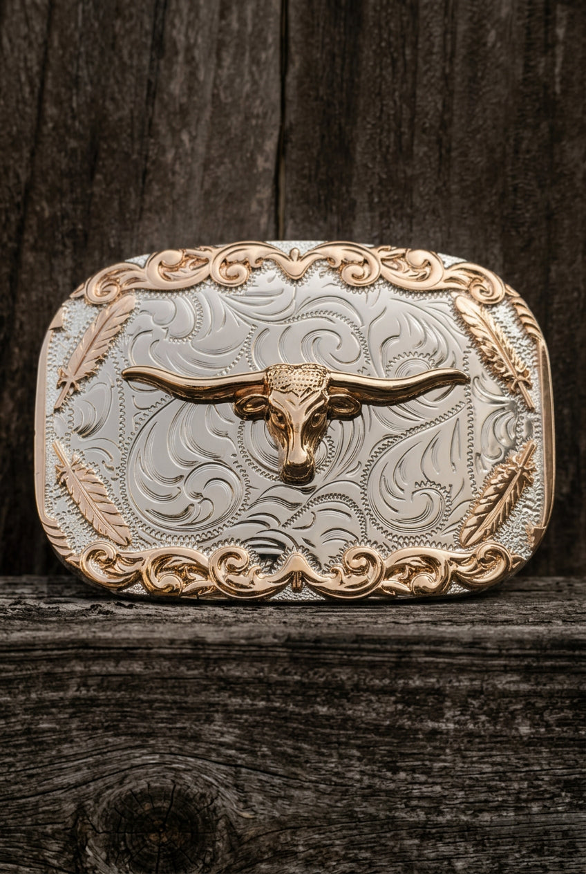 A western-style metal belt buckle featuring a raised longhorn steer motif over an engraved scrollwork design, photographed against a weathered gray wooden surface.