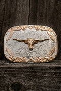 A western-style metal belt buckle featuring a raised longhorn steer motif over an engraved scrollwork design, photographed against a weathered gray wooden surface.